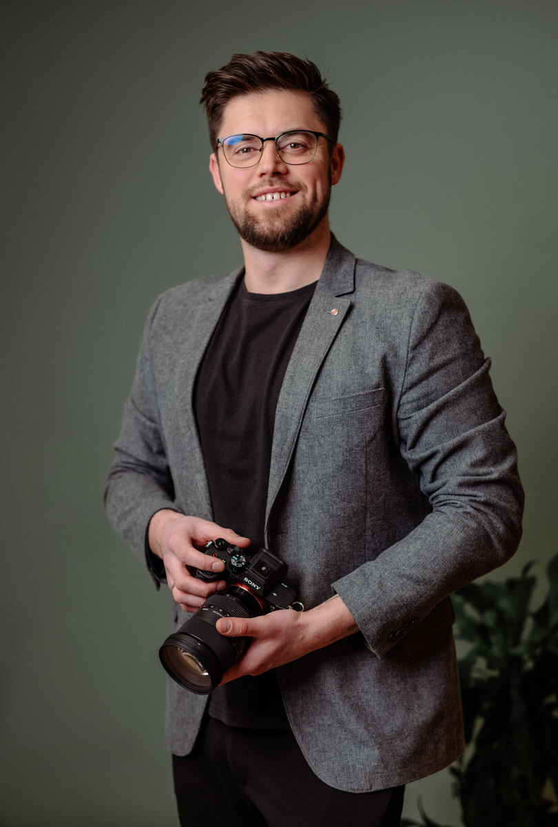 Fotograf Ahaus mit Kamera im Business-Outfit vor grünem Studiohintergrund.